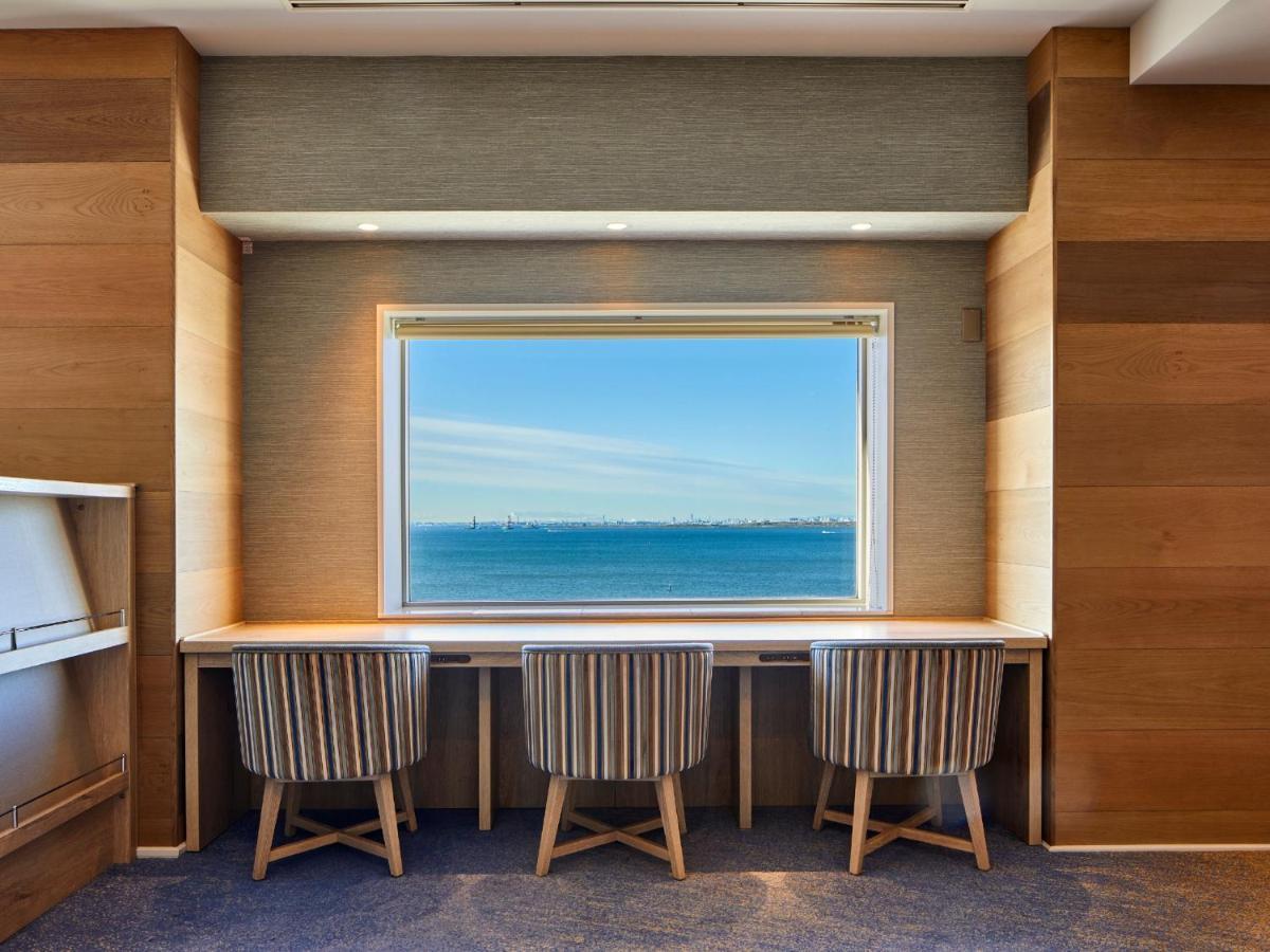 Grand Nikko Tokyo Bay Maihama Ξενοδοχείο Urayasu Εξωτερικό φωτογραφία The photo shows a modern workspace with three striped chairs positioned at a long, white desk. The desk faces a large window with a view of a tranquil body of water and a distant skyline. The walls around the workspace are made of wood paneling, crea
