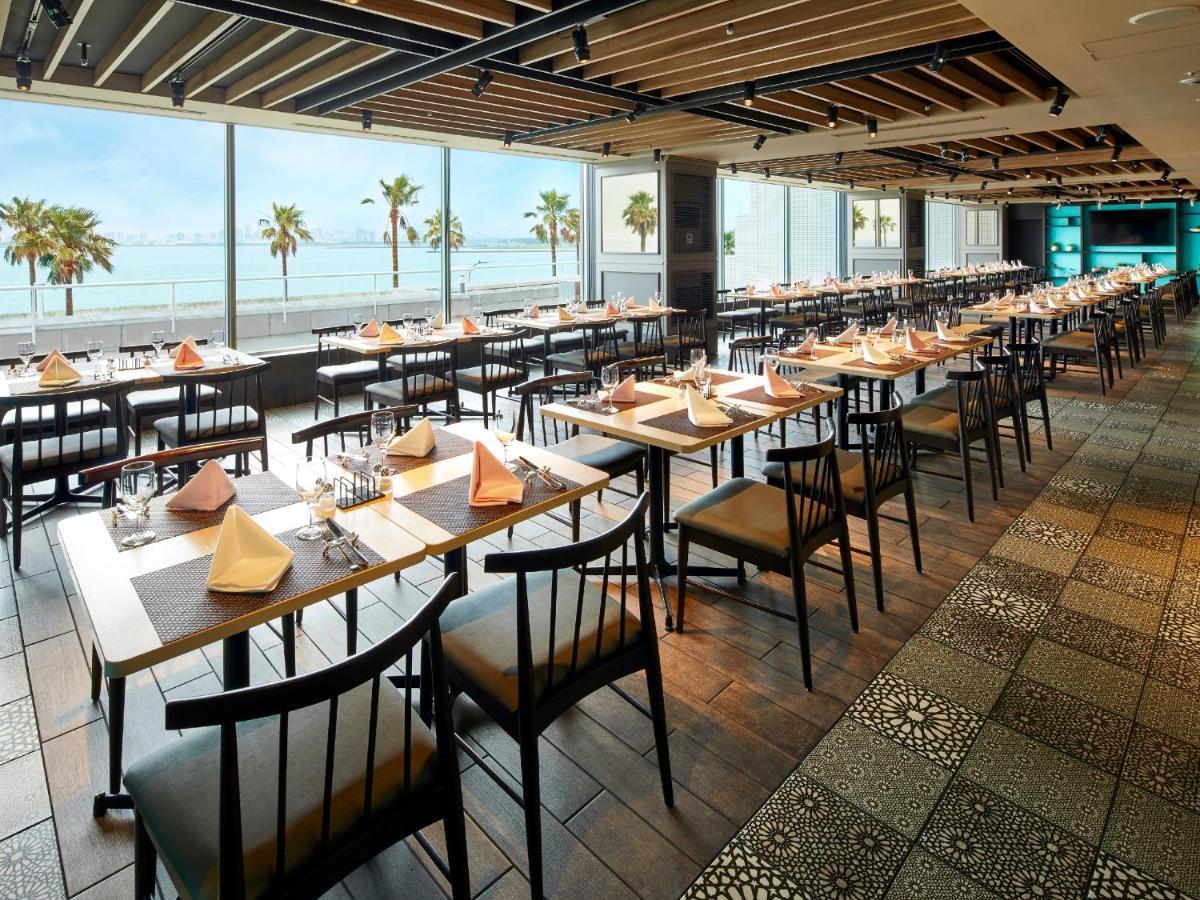 Grand Nikko Tokyo Bay Maihama Ξενοδοχείο Urayasu Εξωτερικό φωτογραφία The photo shows a modern restaurant interior featuring rows of neatly arranged tables and chairs. The large windows provide a scenic view of palm trees and what appears to be a body of water in the background, suggesting a coastal location. Each tabl
