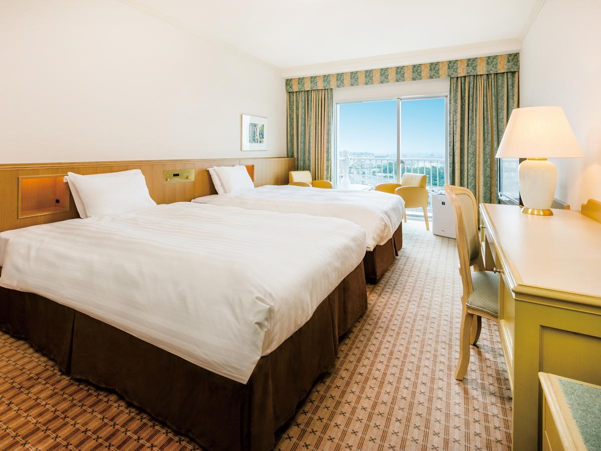 Grand Nikko Tokyo Bay Maihama Ξενοδοχείο Urayasu Εξωτερικό φωτογραφία The photo shows a hotel room featuring two twin beds with white linens and dark brown bed skirts. The room has a light, neutral-colored wall and carpet. There is a window with curtains that let in natural light, offering a view outside. A small desk 
